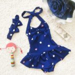 Girl's one-piece swimsuit with blue headband with white dots and a doll next to it on a white carpet