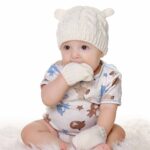 White baby winter bonnet and mittens set with baby with his hand in his mouth and white patterned clothing