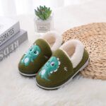 Children's green dinossaure cotton slippers, in front of a plant on a white carpet