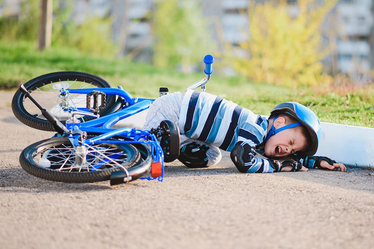 A young child who has fallen off his blue bike. He is crying and wearing a bicycle helmet, knee protectors and black protective gloves