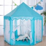 A children's tepee in the shape of a blue princess castle. It stands on the floor in a living room, with a large window behind it. All the walls are made of transparent mosquito netting, and the front curtains are in the open position, with the curtains hanging along the uprights.