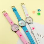 three watches laid flat on a yellow table, one pink, one blue, and one white, they have a cute little cat in the dial and two cat ears above the dial